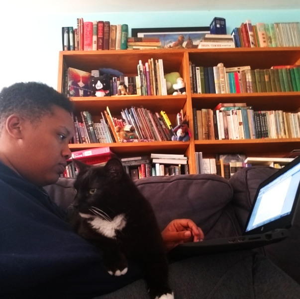 Photo of N. Romaine White on a couch in front of a large bookshelf, a black and white cat and a laptop in her lap.
